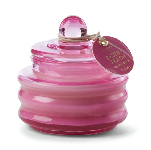 Pink glass jar candle with a visible round brand tag detailing scent of candle "Desert Peach" on a white background. 