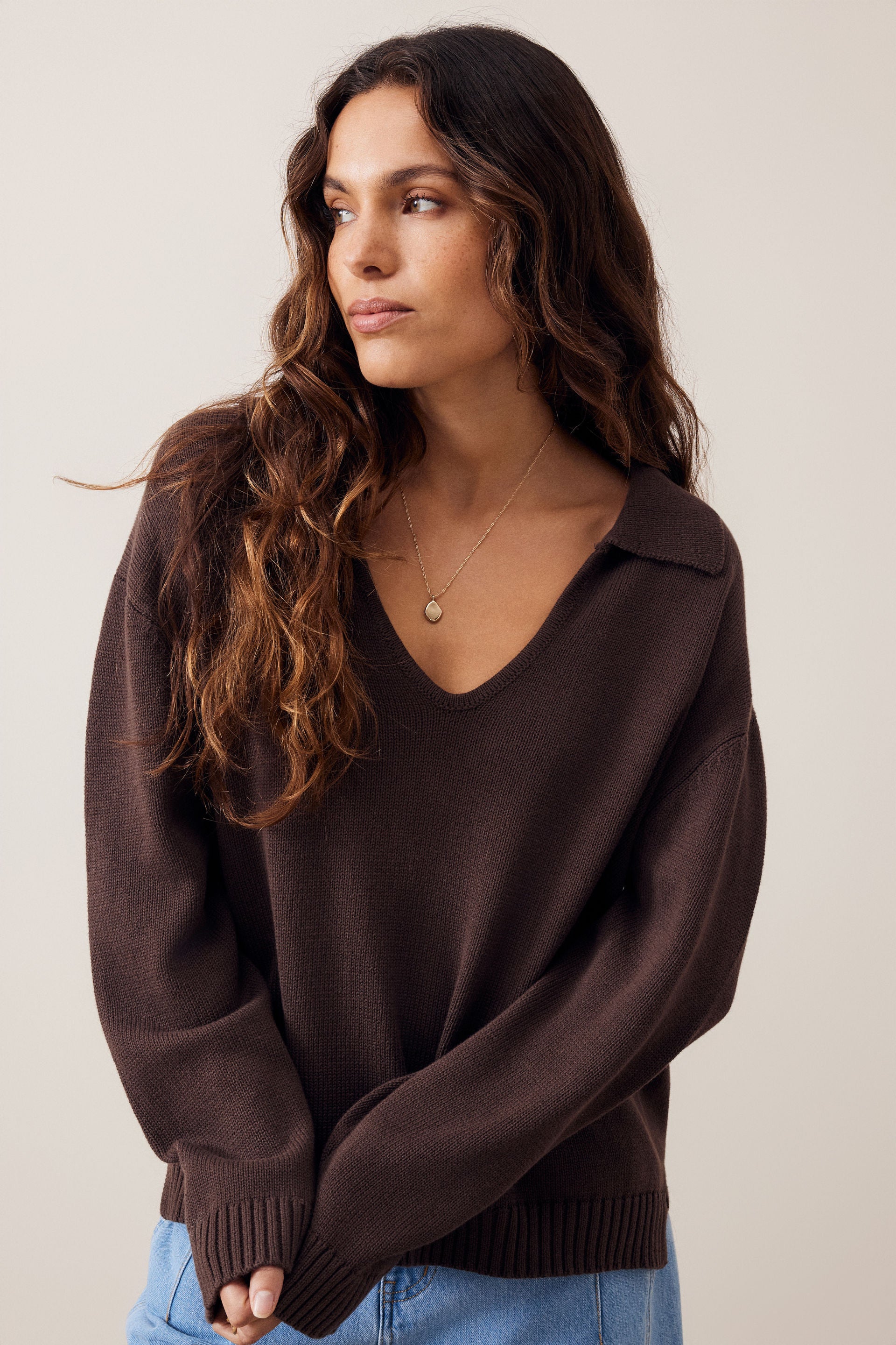 A woman wearing a dark brown v-neck knit sweater with a wide collar and ribbed cuffs, paired with light blue jeans, photographed against a plain light beige background.