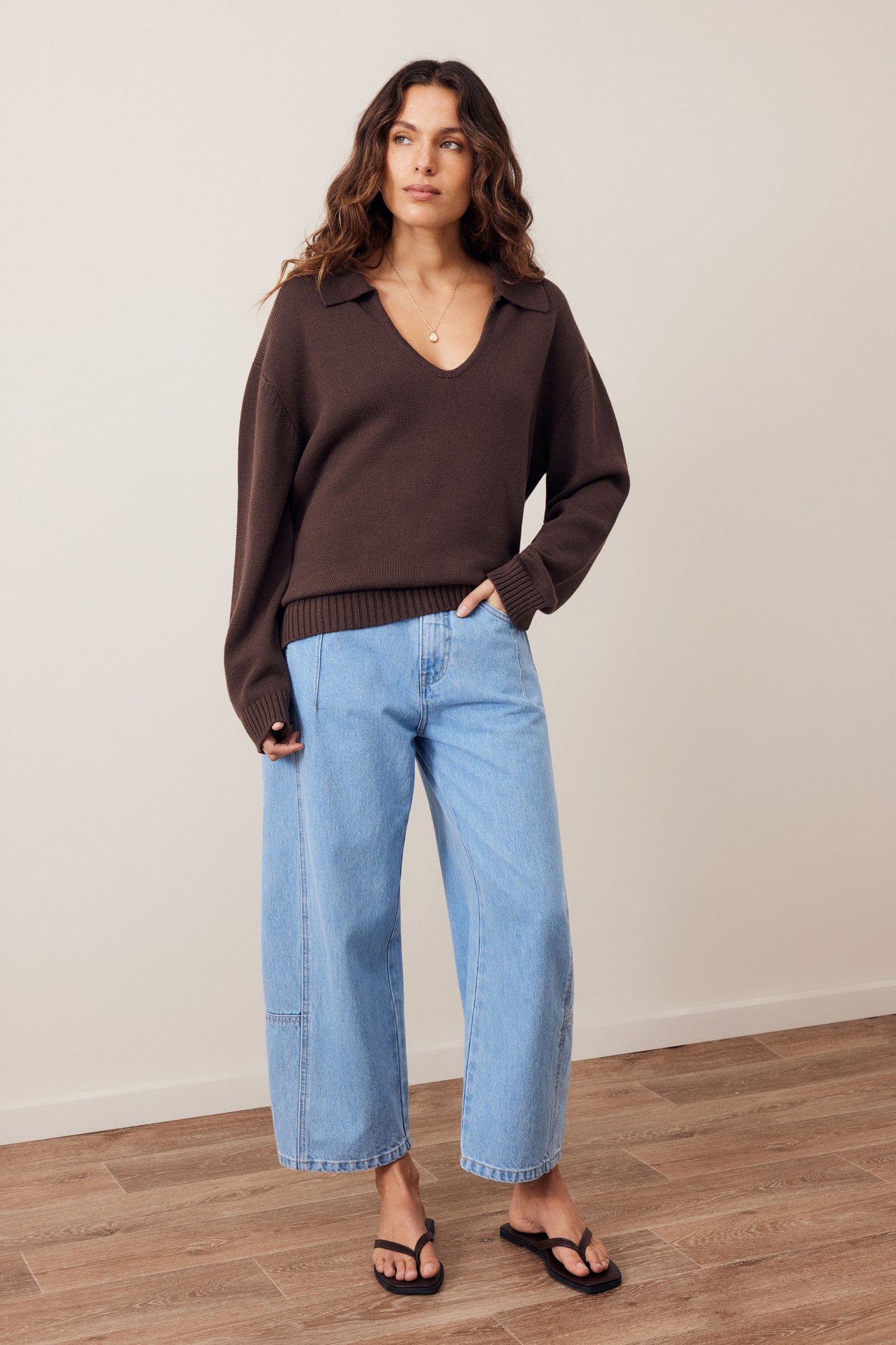 A woman modeling a dark brown long-sleeve knit polo sweater paired with light-wash wide-leg cropped jeans and black flip-flops, standing against a neutral beige wall and wood floor.