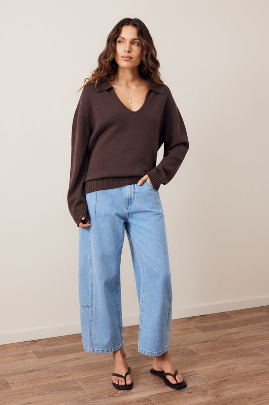 A woman modeling a dark brown long-sleeve knit polo sweater paired with light-wash wide-leg cropped jeans and black flip-flops, standing against a neutral beige wall and wood floor.