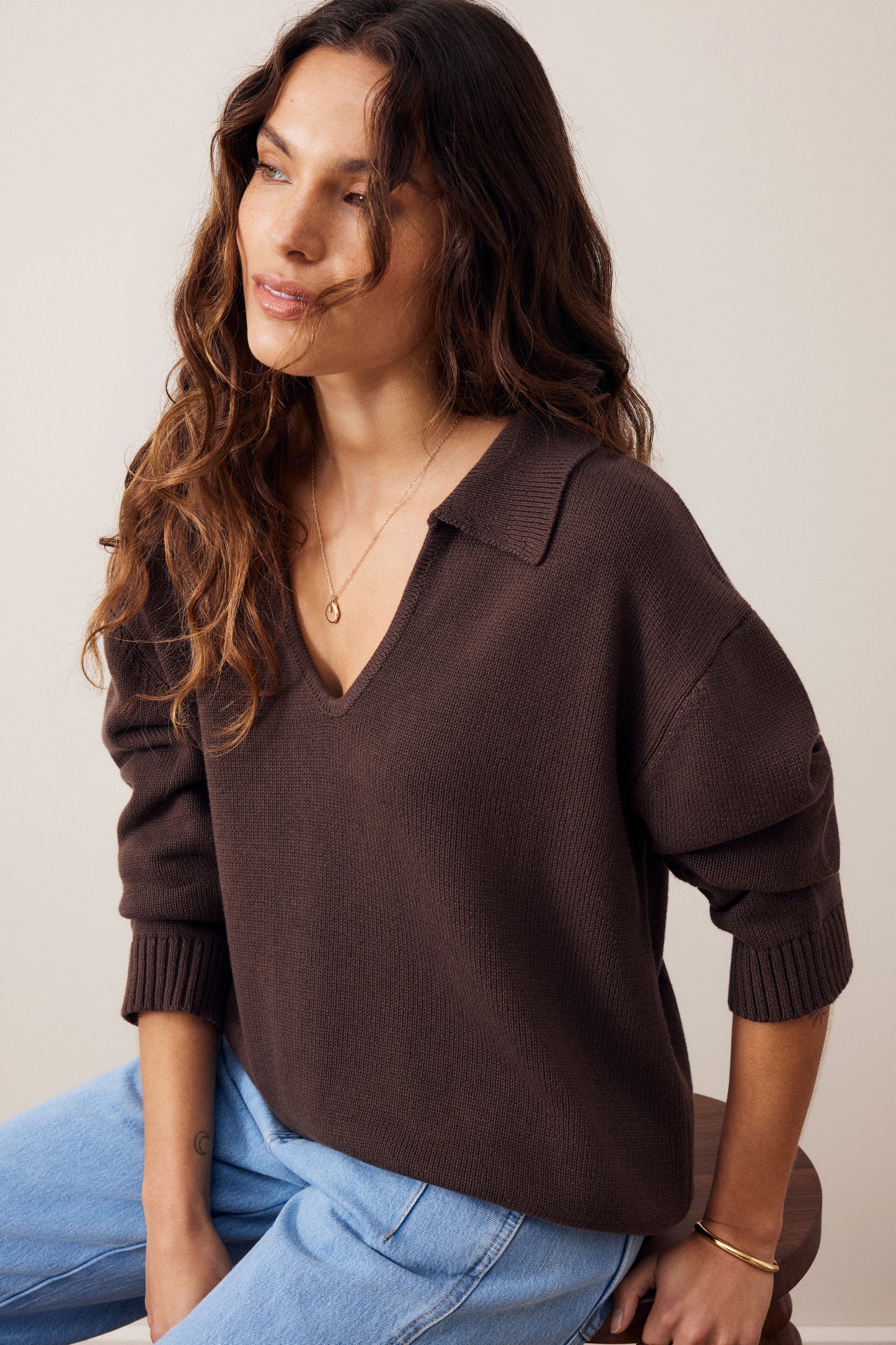 A woman wearing a dark brown ribbed knit sweater with a wide collar and three-quarter sleeves, paired with light blue jeans, photographed against a plain light beige background.