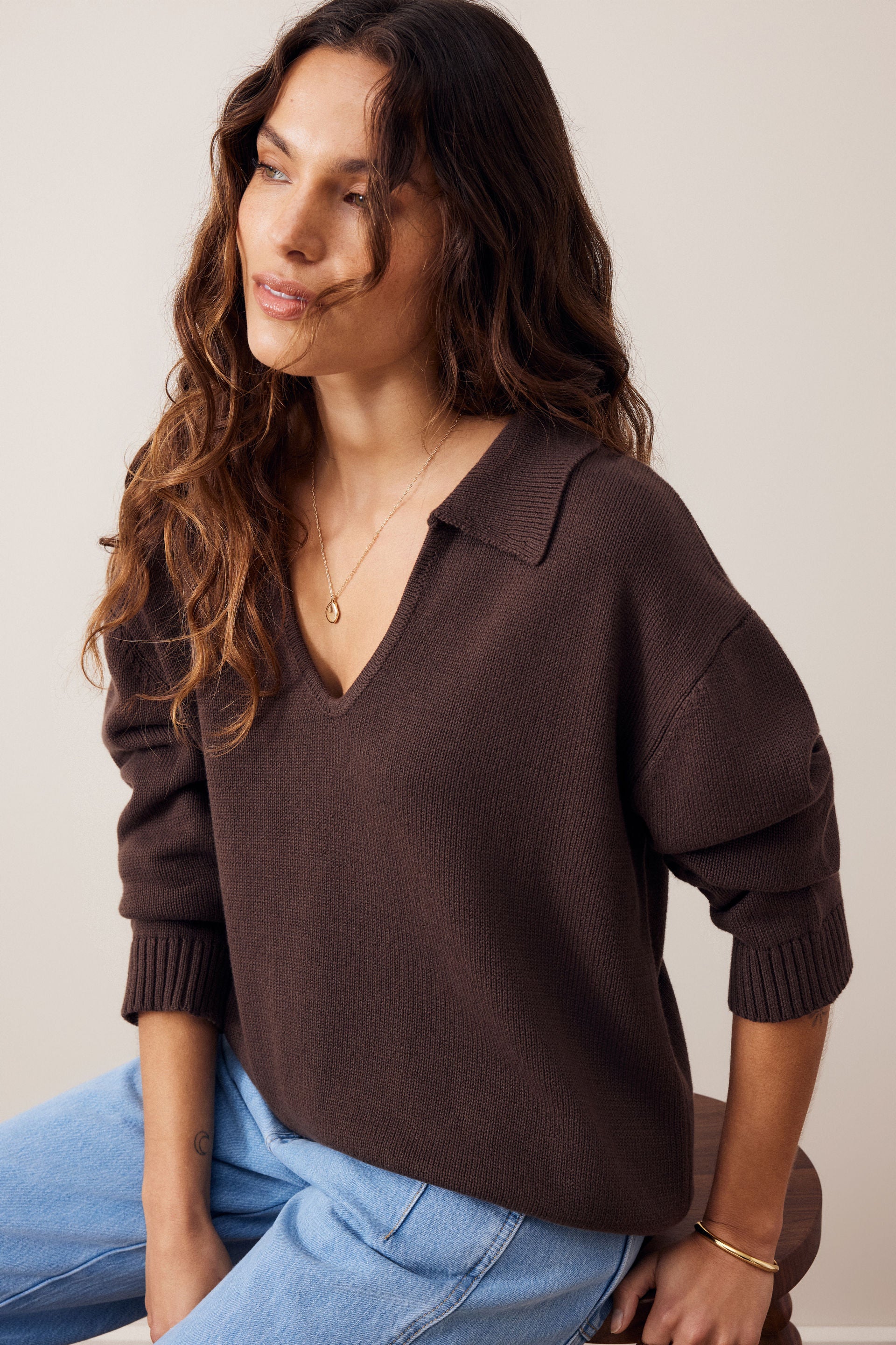 A woman wearing a dark brown ribbed knit sweater with a wide collar and three-quarter sleeves, paired with light blue jeans, photographed against a plain light beige background.