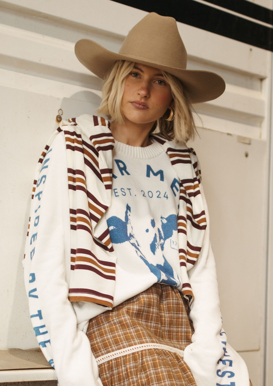 A woman wearing a tan cowboy hat, a white crewneck sweater with brown and rust stripes and blue graphic text, and a brown plaid skirt, posed against a white paneled wall.