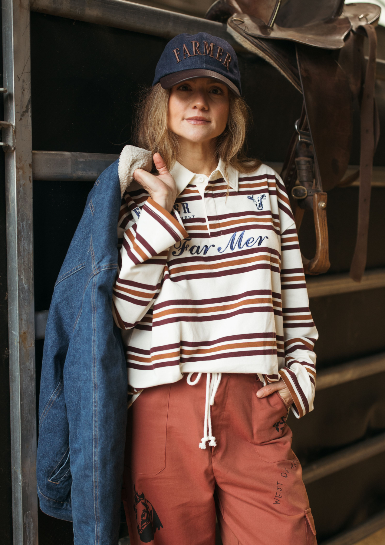 A woman wearing a navy blue 'FARMER' baseball cap, a cream and brown striped long-sleeve polo shirt, rust-colored drawstring pants, and a blue denim jacket, posed against a rustic wooden background with a leather saddle.
