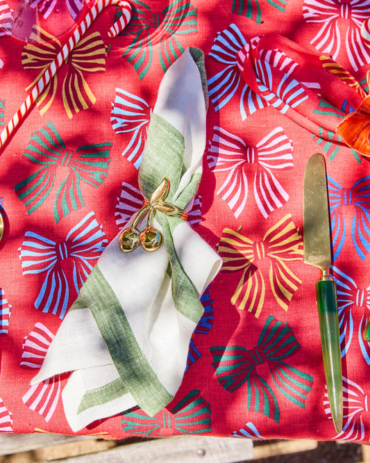 A festive table setting featuring a red tablecloth with a colorful bow pattern, a white and green striped napkin tied with a gold bow, and a green-handled knife.