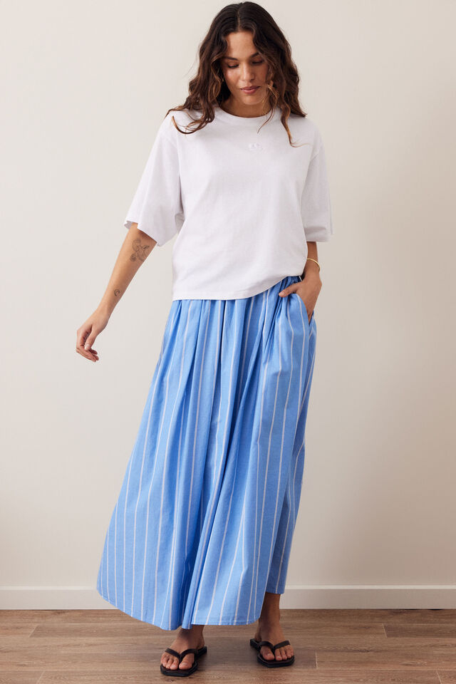 A woman modeling a white short-sleeve t-shirt and light blue wide-leg pleated trousers with thin white vertical stripes, paired with black sandals, standing against a neutral beige wall.