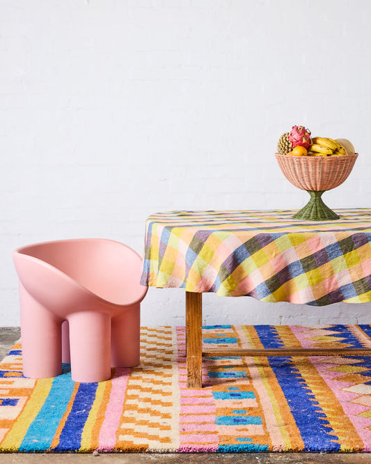 A pink sculptural chair sits on a colorful striped rug next to a table with a yellow, blue, and pink plaid tablecloth and a decorative fruit bowl.
