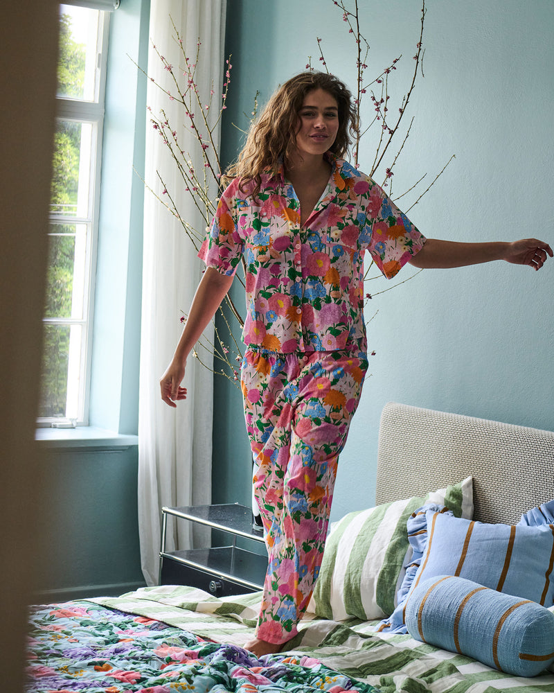 A woman wearing a colorful floral pajama set stands on a bed in a bedroom. The pajama set features a vibrant pink, orange, yellow, and blue floral pattern.