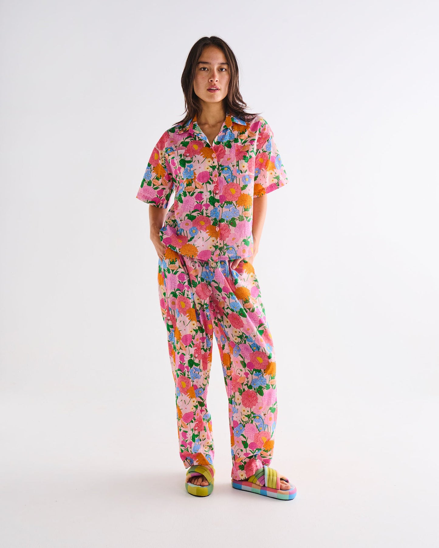 A woman modeling a matching short-sleeve button-up shirt and wide-leg pants pajama set in a vibrant pink floral print, paired with colorful slide sandals, against a plain white studio background.