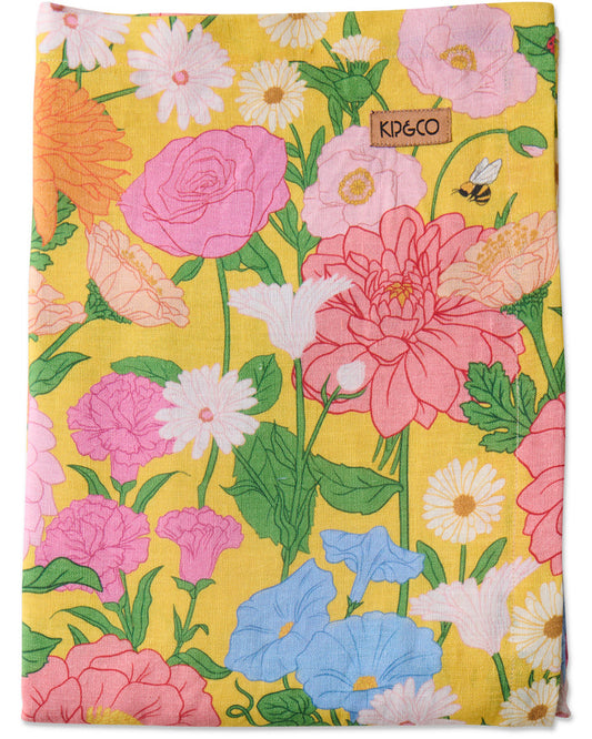 A square fabric item, likely a tea towel or handkerchief, featuring a vibrant floral print on a bright yellow background. The pattern includes large pink and coral roses, white daisies, light blue morning glories, and green leaves.