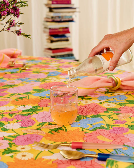 A hand pours a clear liquid from a glass bottle into a clear glass on a brightly patterned floral tablecloth. A pink napkin, gold spoon, fork, and other tableware are visible, with a blurred background of books and curtains.