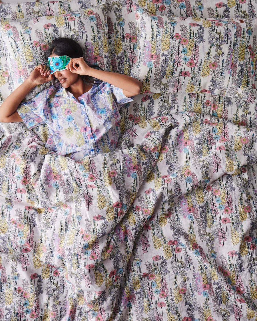 Person lying in bed with a sleeping mask on a foxglove floral patterned bedspread, person wears matching pyjamas to bedspread,. 