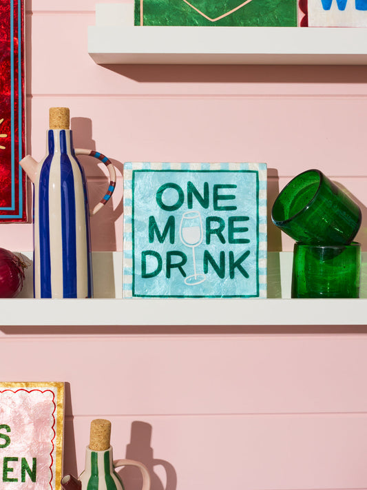 Decorative sign with 'One More Drink' text on a shelf with bottles and glasses.