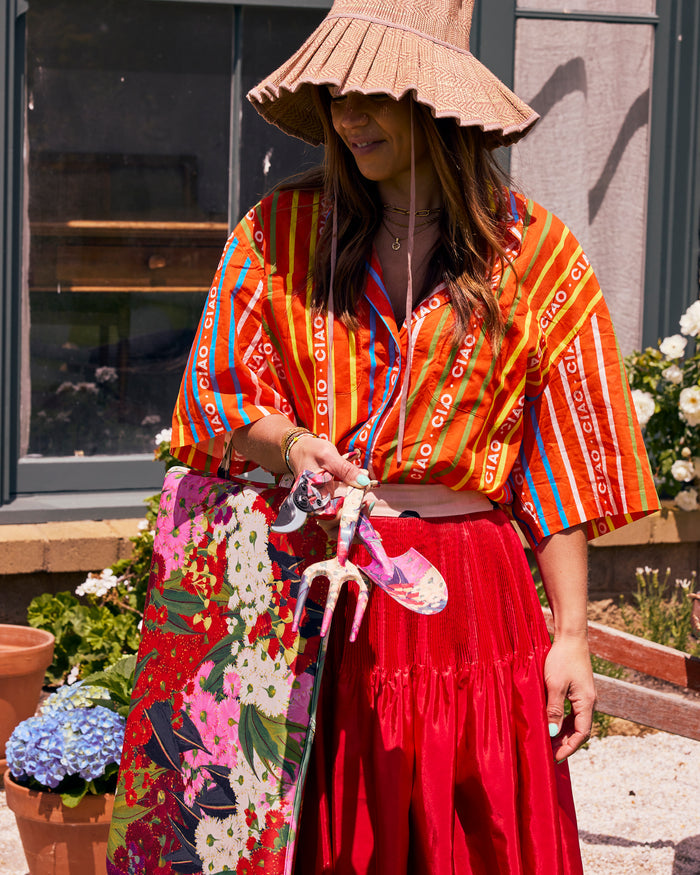 A woman wearing a bohemian-style outfit featuring a vibrant orange and red patterned blouse, a bright red skirt, and a large woven straw hat, standing outdoors in front of a building.