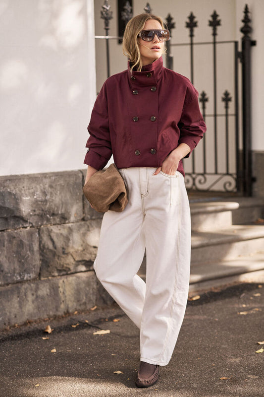Woman in burgundy jacket and white pants standing outdoors near a metal gate.