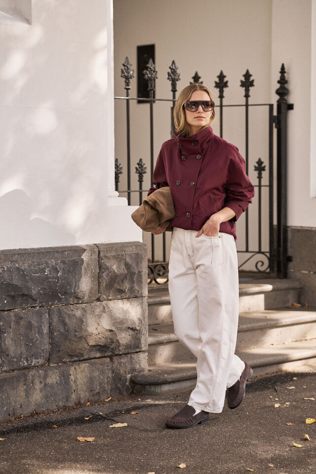 A woman wearing a double-breasted burgundy jacket, white trousers, and dark loafers stands on outdoor stone steps next to a white building with a black wrought-iron gate.