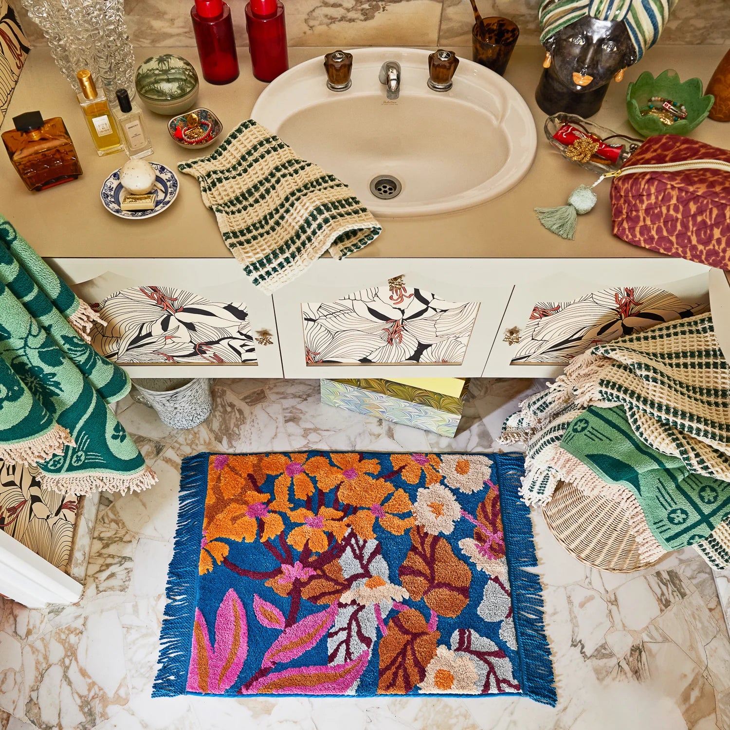 Bathroom with a colorful bath mat on the floor, towels hanging, and various cosmetics on the counter.
