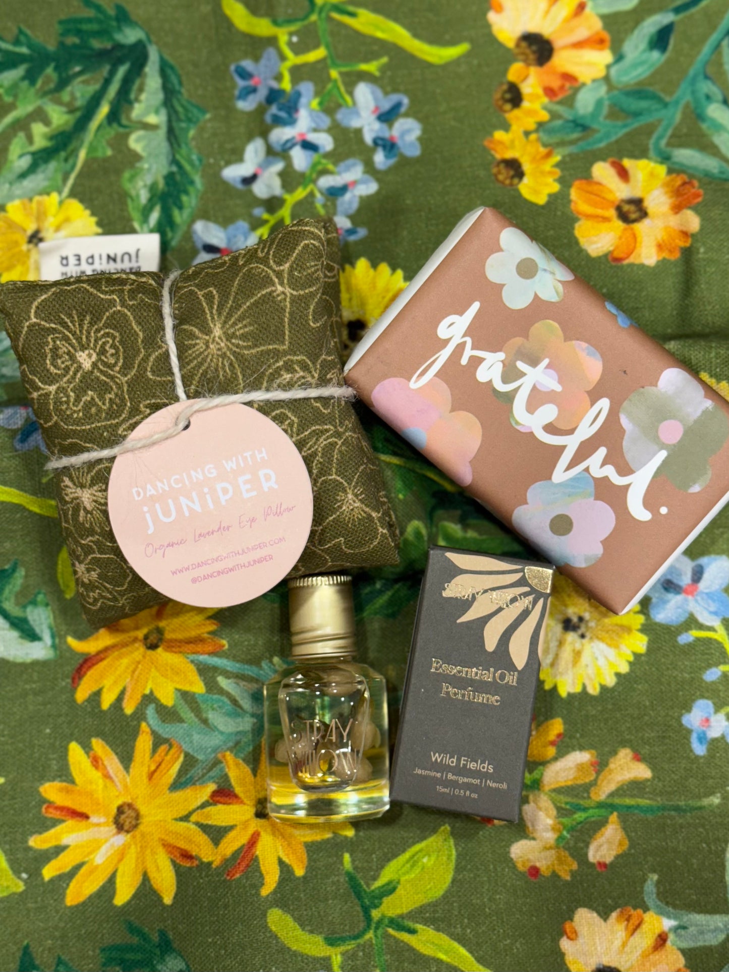 Essential Oil perfume roller bottle from Stray Willow, wrapped soap bar with grateful written on front and flower details, and small eye pillow with khaki green floral fabric from Australian made and designed brand Dancing With Juniper on a floral fabric background.