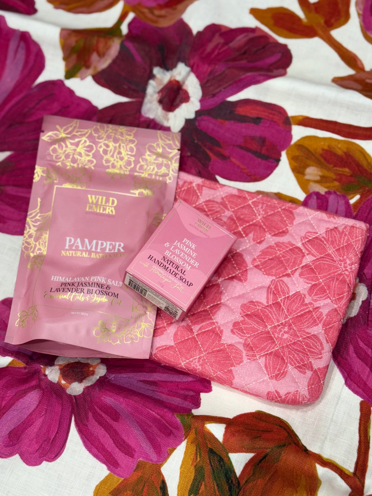 Pink floral cosmetic bag, pamper natural bath salts and natural soap bar from Wild Emery laid on a floral-patterned surface