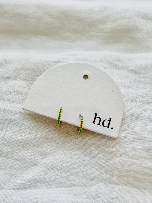 Pair of green and gold earrings on a white card with 'hd.' text, placed on a textured beige surface.