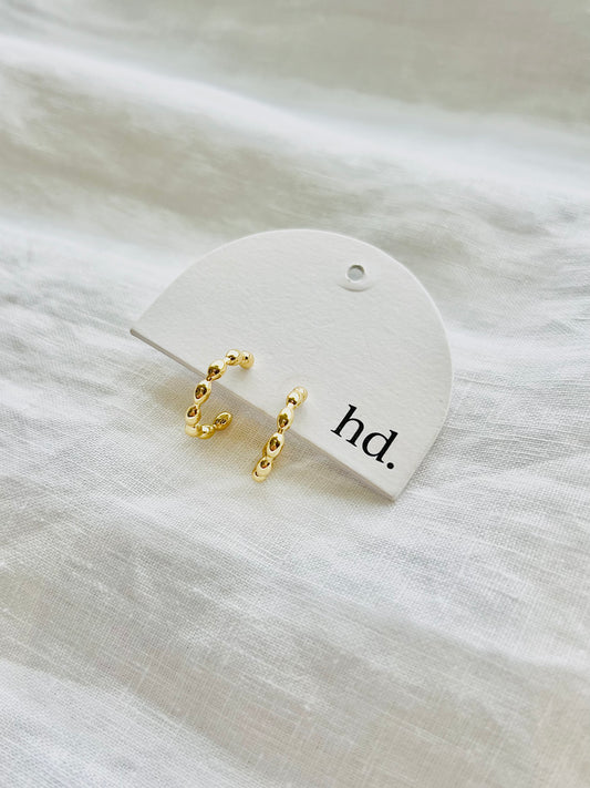 Gold hoop earrings on a white card with 'hd.' text, placed on a textured white surface.