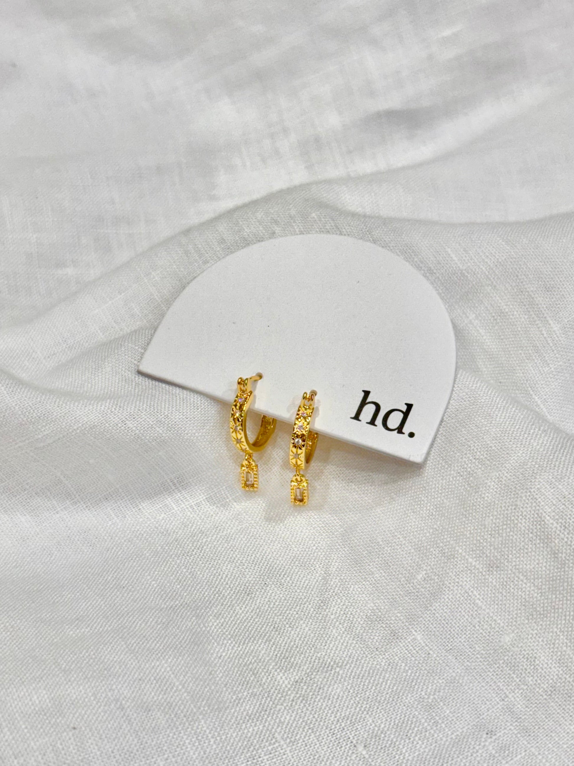 Gold earrings on a white card with 'hd.' text, placed on a white fabric background