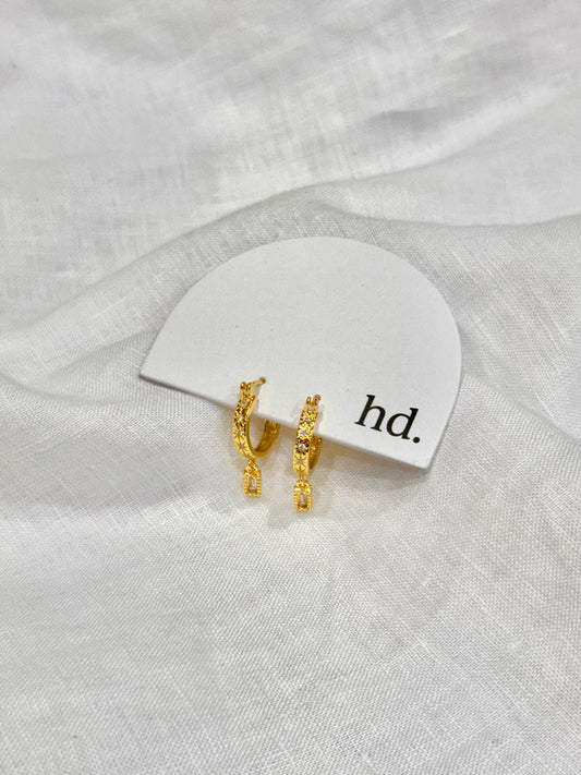 Gold earrings on a white card with 'hd.' text, placed on a white fabric background