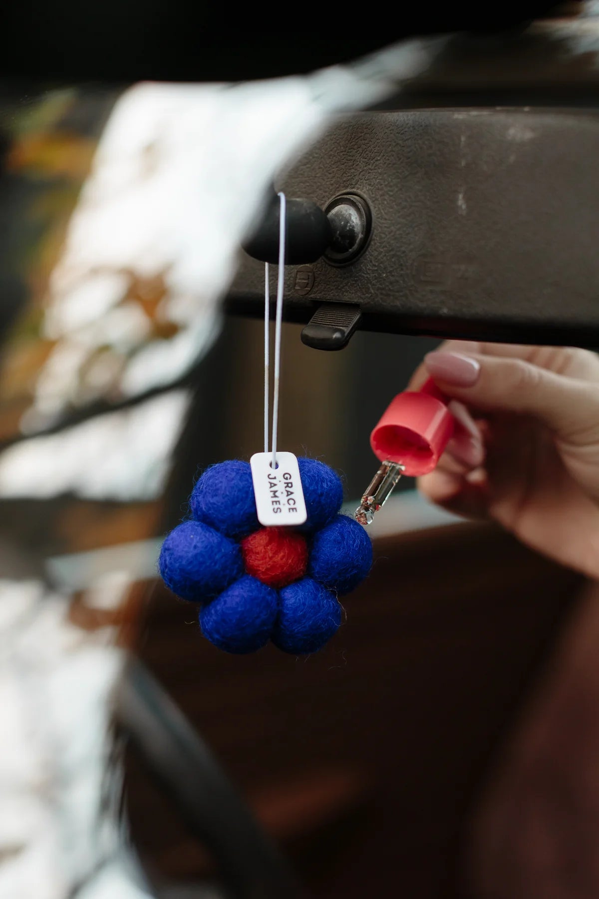 Blue and red felt flower air freshener hanging from mirror inside car with blurred natural background. Hand holding dropper of fragrance dropping oil on air freshener. 