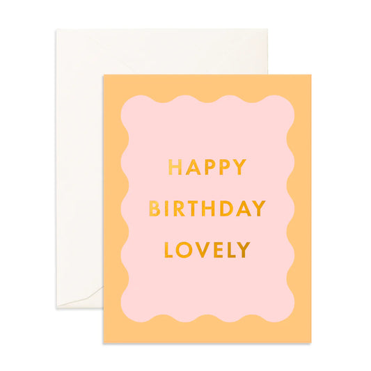 A greeting card with 'HAPPY BIRTHDAY LOVELY' written in gold on a pink background, with a scalloped border and a white envelope.