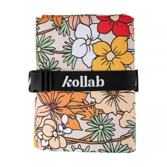 A rolled-up picnic mat, featuring a retro floral pattern in orange, yellow, red, green, and white on a beige background, secured with a black strap branded with 'kollab' in white text.