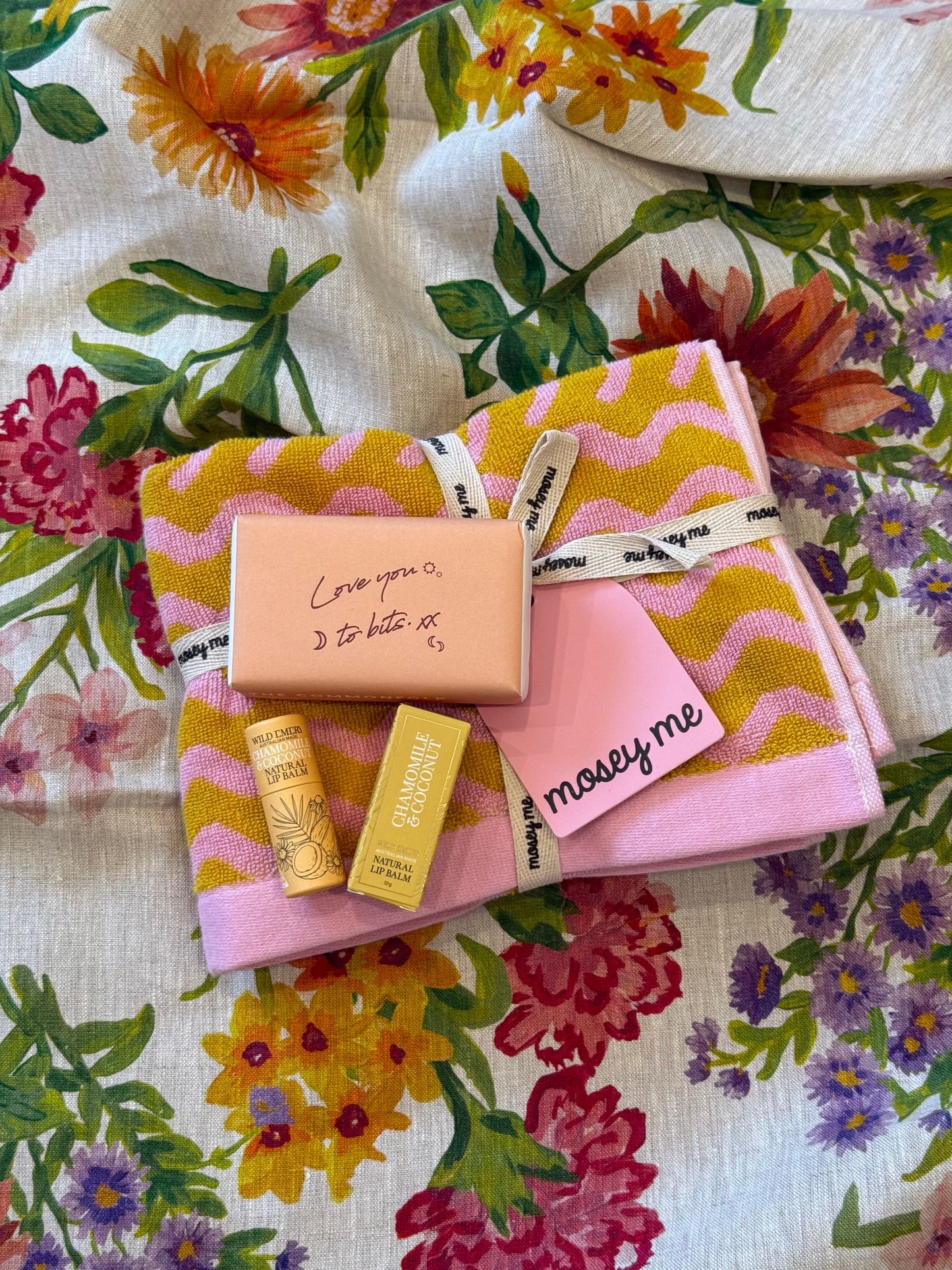 Yellow and Pink zig zag pattern hand towel from Mosey and Me, sat on top of hand towel is a wrapped soap bar with saying "Love You To Bits" written on packaging and Wild Emery Lip Balm in Chamomile + Coconut scent with yellow packaging on a floral fabric background.