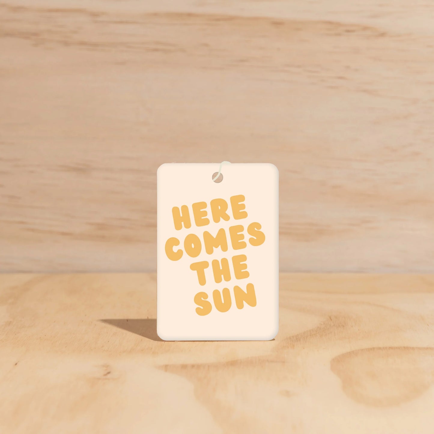 Rectangular car freshener with 'HERE COMES THE SUN' text on a wooden surface