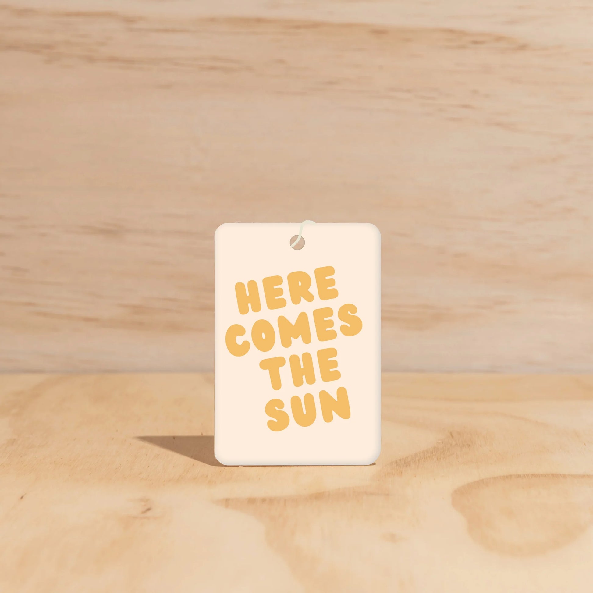 Rectangular car freshener with 'HERE COMES THE SUN' text on a wooden surface