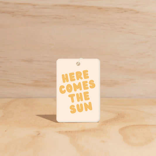 Rectangular car freshener with 'HERE COMES THE SUN' text on a wooden surface