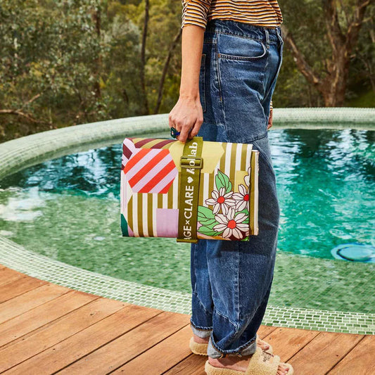 A person standing by a swimming pool holds a colorful clutch bag. The bag has a multicolor geometric and floral print with a green strap that reads 'SAGE X CLARE x KOLLAB'.