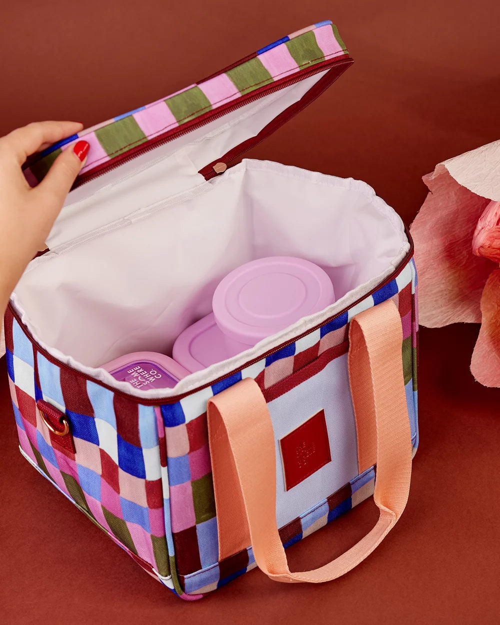 A square soft-sided lunch bag with a multicolor plaid pattern, shown open with a hand lifting the lid. Inside are two pink reusable containers.
