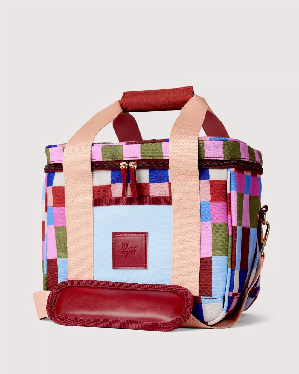 A square soft-sided cooler bag with a colorful patchwork pattern in pink, blue, green, and brown, featuring light beige handles and a dark red leather handle, front patch, and bottom pad, set against a light gray background.