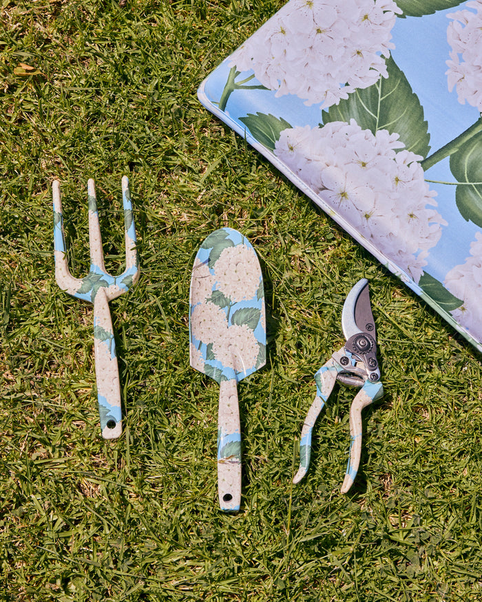 Three garden hand tools with a blue, white, and green hydrangea pattern are arranged on green grass next to a matching fabric pad. The tools include a three-prong garden fork, a trowel, and pruning shears.