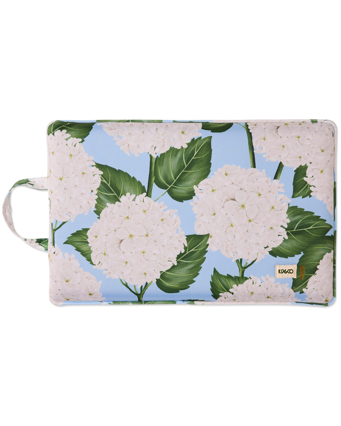 A rectangular soft pouch with rounded corners featuring a watercolor-style floral print of large white hydrangeas with green leaves on a light blue background. A small tan square label with black text is visible near the lower right corner, and a white carrying loop is attached to the left side.