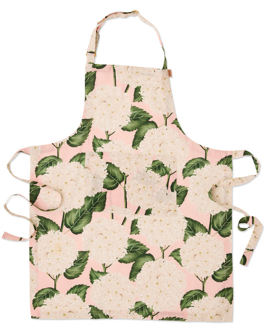 A floral apron with a pink background and a repeating pattern of large white hydrangea flowers and green leaves. It features a neck strap, waist ties, and side ties, shown flat against a white background.