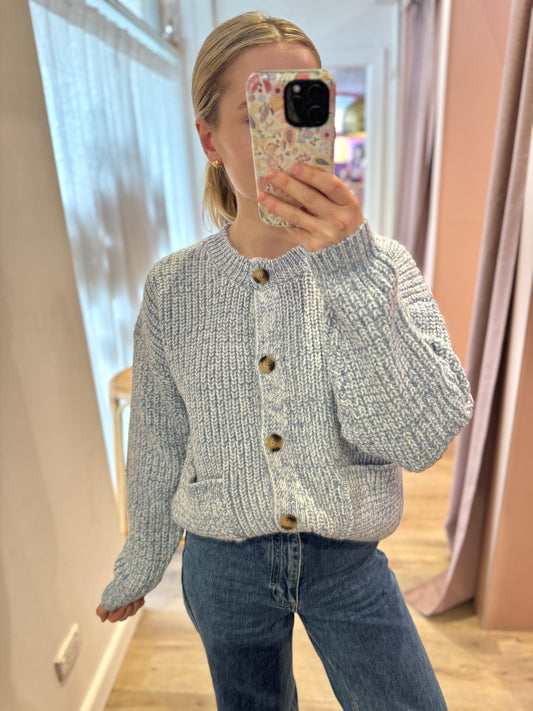 Person wearing a gray knitted cardigan with brown buttons in a room with pink curtains.