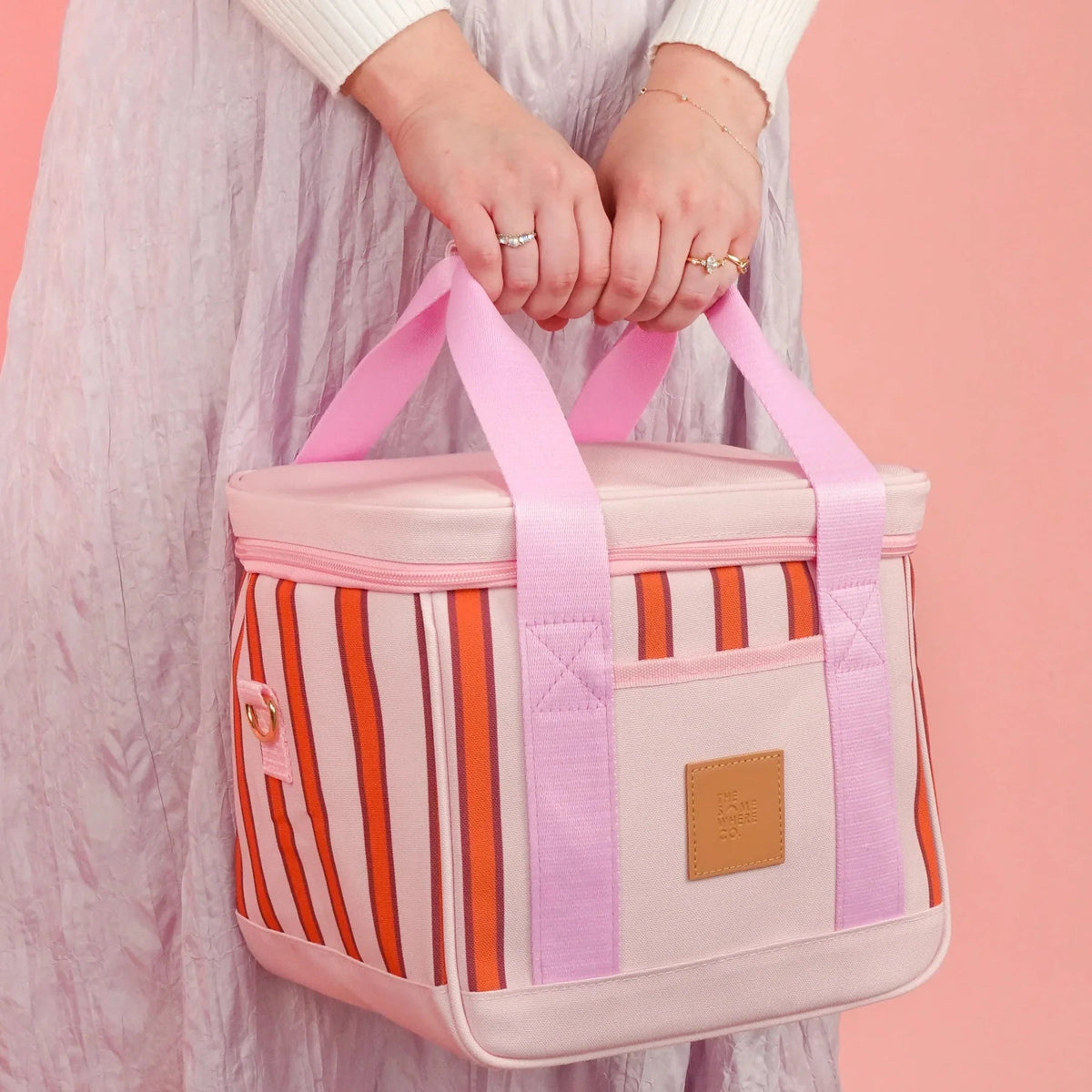 A person in a light pink dress holds a square insulated tote bag with pink handles and pink, orange, and white vertical stripes. The bag has a front pocket with a tan patch and a small pink tag, set against a solid pink background.