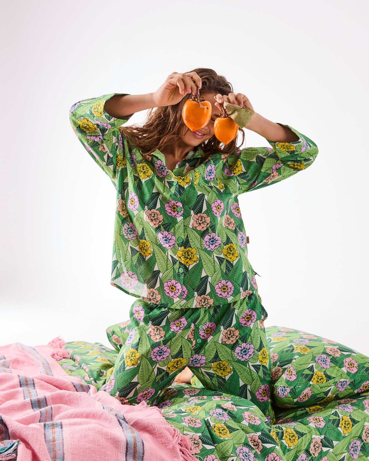 A woman wearing a green floral pajama set sits on a white studio floor, holding two orange peels over her eyes. The pajamas feature a colorful botanical print with pink, yellow, and purple flowers and green leaves.