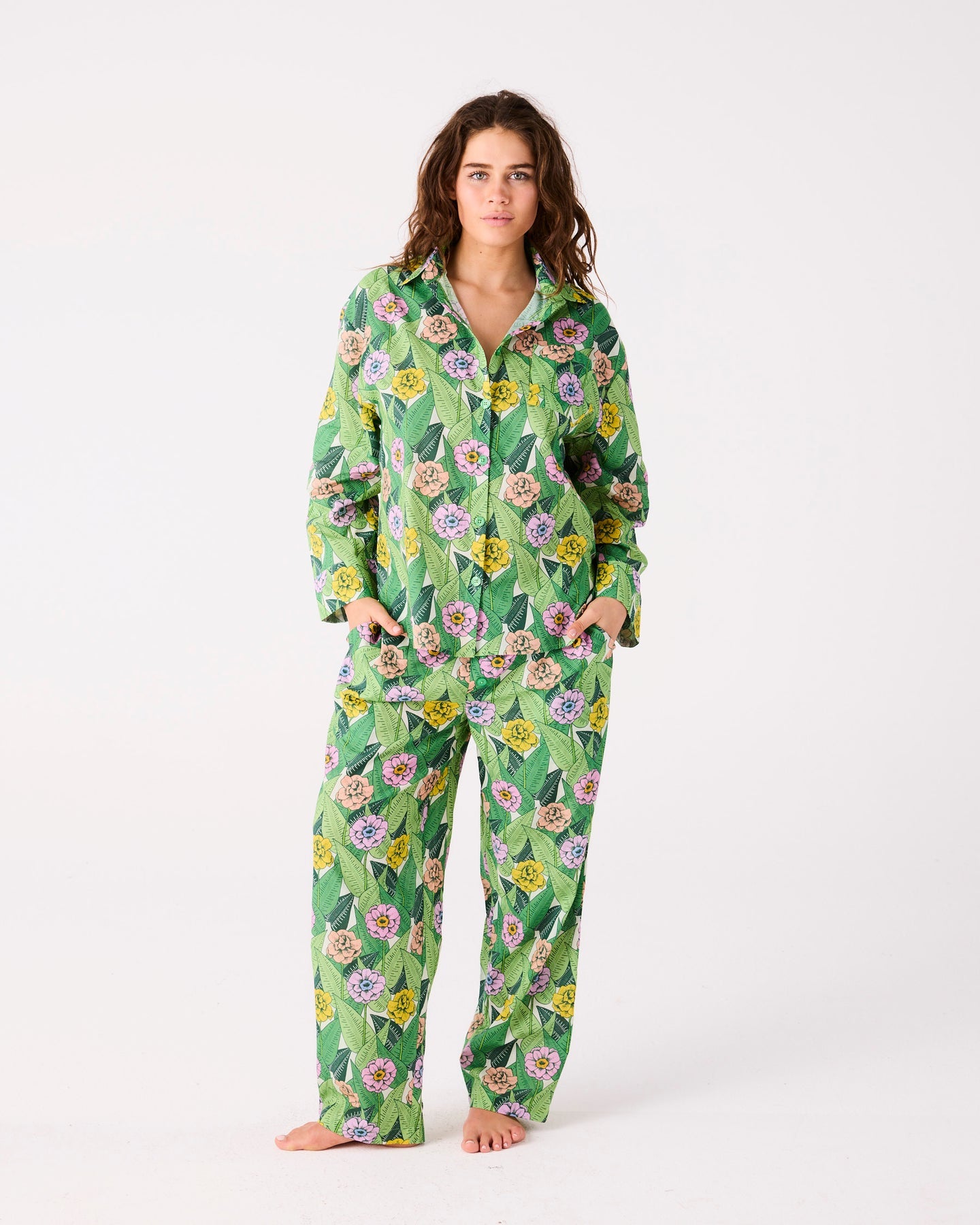 A woman modeling a matching two-piece pajama set with a green tropical floral print, featuring a long-sleeve button-up top and wide-leg pants, against a plain white studio background.