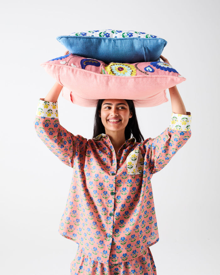 A woman wearing pink floral pajamas balances a stack of two colorful pillows on her head against a plain light gray background.