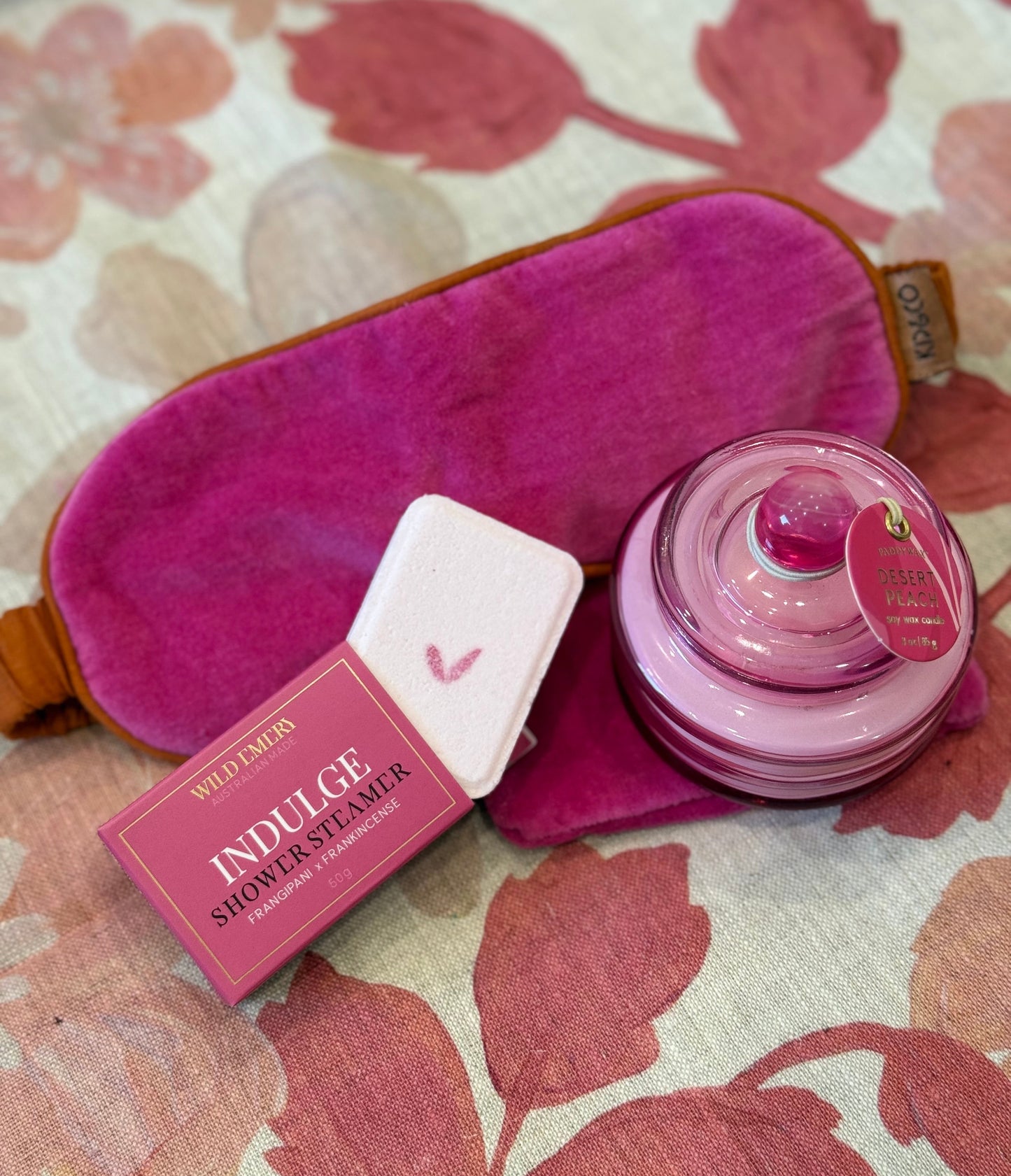 Hot pink velvet eye mask, shower steamer from Wild Emery with boxed packaging, and pink jar candle on a floral-patterned surface