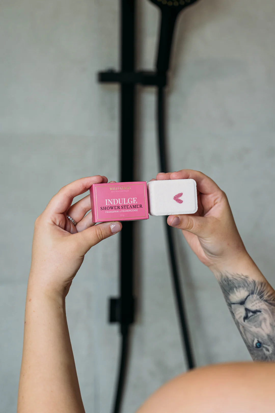 Person holding shower steamer with pink heart, and packaging detailing name "Indulge" in a bathroom setting.