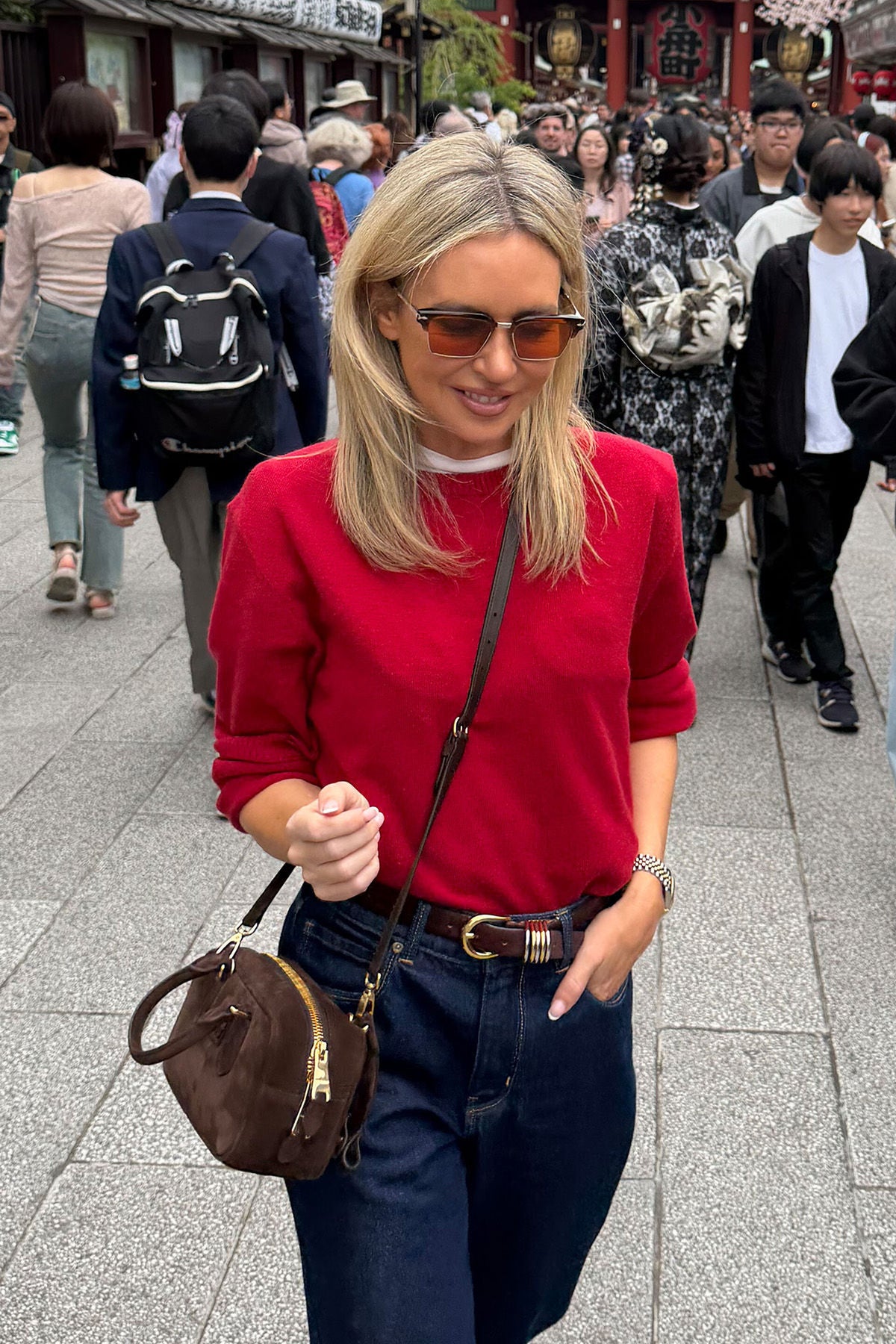 Woman in a red sweater and sunglasses walking on a busy street.