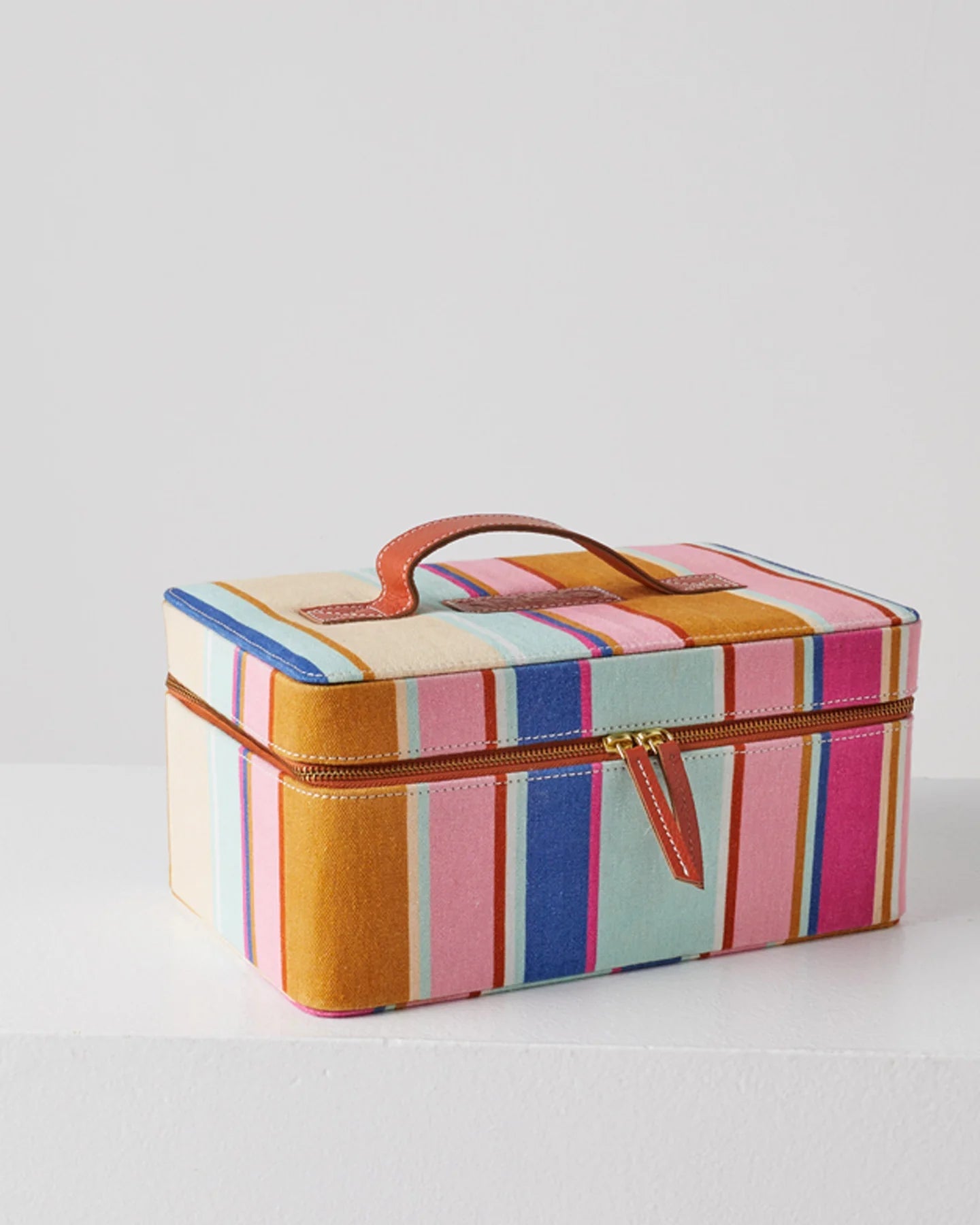 Closed toiletry case shows brown leather zip handles and top handle. Stripe pattern fabric outer includes blue, pink, mustard, and small red stripe. 
