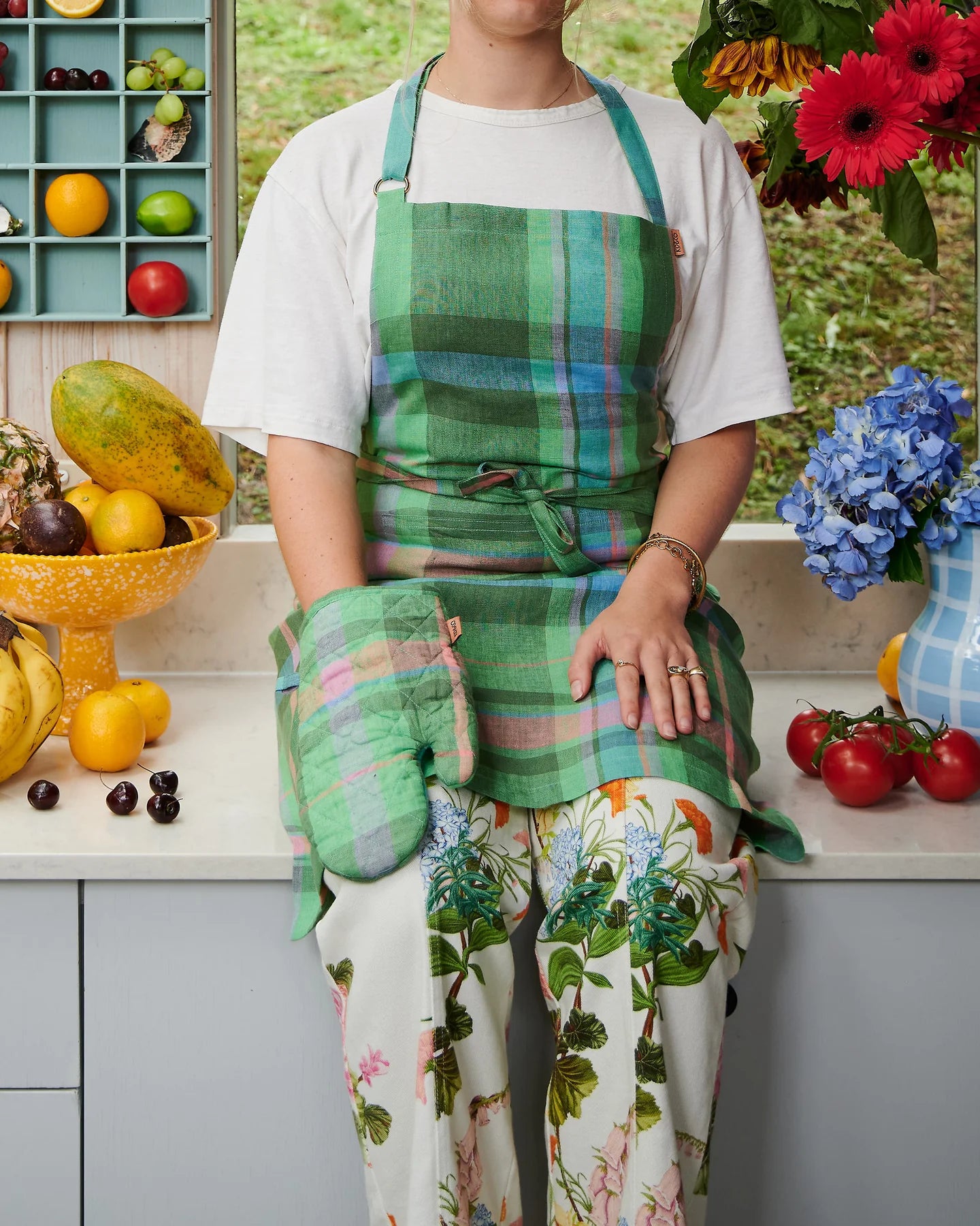 Woman sits on kitchen bench surrounded by fruit and jug filled with flowers, she wears apron and matching oven mitt in green base, with blue pink and lilac tartan pattern oven mitt. 